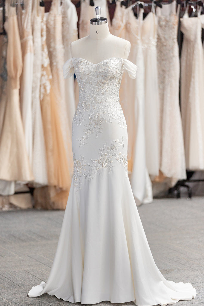 A beautiful off-the-shoulder wedding dress made of crepe fabric with a fit-and-flare silhouette. The dress is adorned with delicate embroidered lace detailing, featuring a sweetheart neckline and flattering off-the-shoulder sleeves.